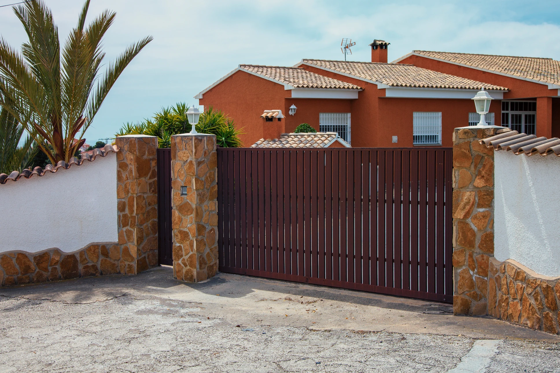 Extérieur résidentiel avec portail coulissant en lames verticales bois foncé, intégré dans clôture mixte pierre naturelle et crépi blanc