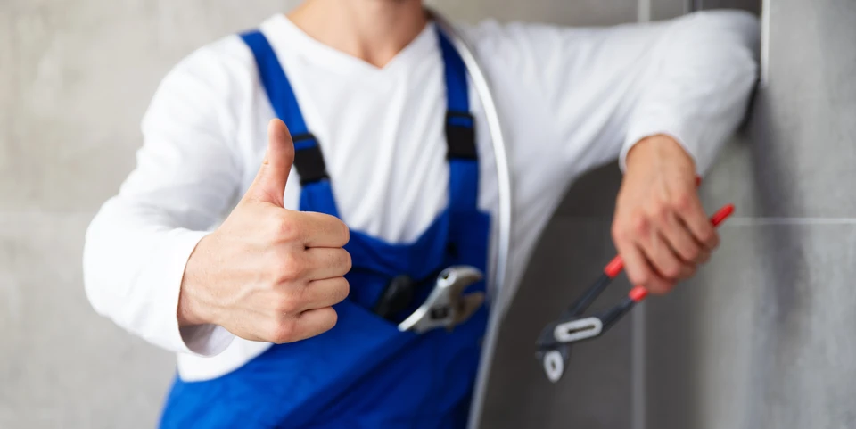 Artisan en salopette bleue et pull blanc fait un geste de pouce levé tout en tenant un tournevis dans un environnement intérieur résidentiel près d'une porte blanche